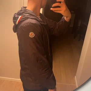 Mörkgrå vindjacka från Moncler med huva - Svart vindjacka från Moncler med klassisk logga på ärmen och huva med snygga röda och vita ränder. Jackan har dragkedja framtill, två fickor med knappar och elastiska muddar vid ärmslut. Perfekt för dig som vill ha en stilren och sportig look.