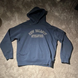 Blå hoodie från New Balance Athletics - Säljer en blå hoodie från New Balance Athletics med tryck på bröstet. Tröjan har huva, känguruficka och långa ärmar. Perfekt för dig som gillar sportig och avslappnad stil.