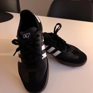 Adidas Samba svarta sneakers - Adidas Samba sneakers i svart skinn med vita detaljer och klassiska tre ränder på sidorna. Brun gummisula, rund tå och snörning framtill. Endast använda fåtal gånger!