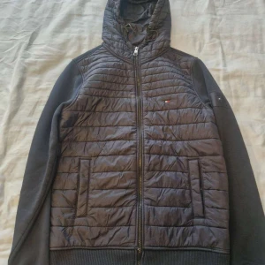 Mörkblå jacka från Tommy Hilfiger - Svart pufferjacka från Tommy Hilfiger med quiltad framsida och huva med snörning. Jackan har dragkedja, ribbade muddar och mjukt fleecefoder på insidan. Små logodetaljer på bröstet och armen. Perfekt för kyliga dagar.
