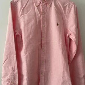 Rosa skjorta från Ralph Lauren - Klassisk rosa skjorta från Ralph Lauren i slim fit-modell. Skjortan har button down-krage, långa ärmar och broderad logga på bröstet. Tillverkad i mjuk bomull och stängs med vita knappar framtill.