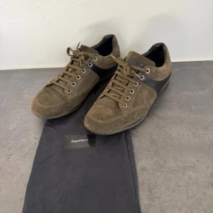 Bruna/Khaki sneakers från Zegna i mocka - Snygga sneakers från Zegna i brun/khaki mocka med svarta skinnpaneler på sidorna. Skorna har rund tå, snörning med metallöljetter och platt sula. Perfekta för dig som gillar en stilren och sportig look med premiumkänsla.
