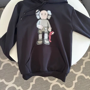 Svart hoodie med KAWS-tryck - Svart hoodie med stor KAWS-figur tryckt på framsidan. Tröjan har huva med snörning och en klassisk känguruficka. Materialet känns mjukt och skönt, perfekt för chill dagar. Snygg streetwear-vibe och coolt motiv i grått, vitt och rött.