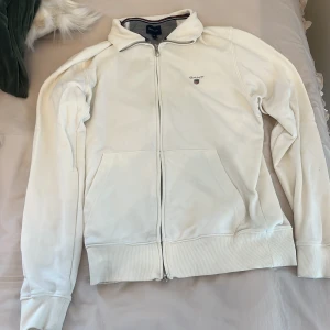 Vit ziptröja från Gant i bomull - Säljer en stilren vit ziptröja från Gant med broderad logga på bröstet. Tröjan har hel dragkedja, ribbade muddar och krage med snygg randig detalj på insidan. Tillverkad i mjuk bomull och har två fickor framtill.