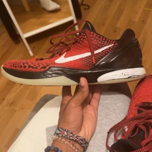 Nike Kobe 6 Protro röd/svart sneakers - Nike Kobe 6 Protro sneakers med röd och svart reptil-inspirerat mönster, vit swoosh och snörning. Skorna har låg profil, platt sula och detaljer i syntetmaterial. Perfekt för dig som gillar basket och vill sticka ut med en unik look.