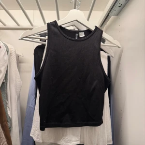 Svart ribbad croppad linnetopp - Svart croppad linnetopp från H&M med ribbad struktur. Toppen är ärmlös och har en rundad halsringning. Perfekt för att styla med jeans eller kjol för en clean och enkel look.