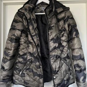 Camouflage pufferjacka - Pufferjacka i grön och svart camouflage. Jackan har huva, dragkedja framtill och två dragkedjefickor. Tillverkad i 100% polyester. Jackan är mer grön än på bild. Storlek 36/38. 