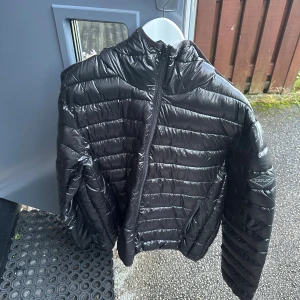 Svart pufferjacka från Replay - Säljer en svart pufferjacka från Replay med glansig finish och horisontella sömmar. Jackan har hög krage, huva och dragkedja framtill samt Replay-logga på ärmen. Perfekt för kyliga dagar och riktigt snygg till streetwear. 