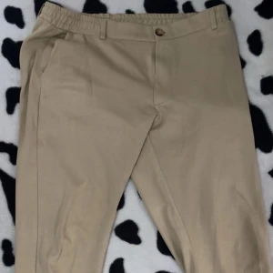Beige chinos med elastisk midja - Snygga beige chinos med elastisk midja och knappstängning. Byxorna har klassiska sidfickor och bälteshällor, vilket gör dem både stilrena och bekväma. Materialet känns lätt och passar perfekt för en chill och clean look.