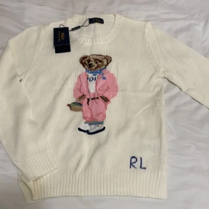 Vit stickad tröja Polo Ralph Lauren - Vit stickad tröja från Polo Ralph Lauren med ikonisk björn broderad på framsidan i rosa outfit och blå scarf. RL-broderi nere till höger. Rund hals och ribbade muddar. Materialmix med merinoull, polyester, akryl och nylon. Perfekt för dig som gillar statement-plagg.