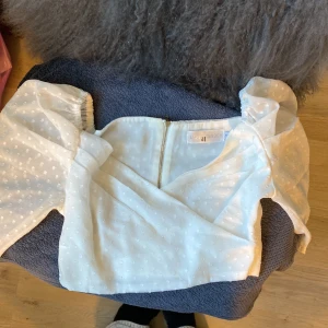 Vit blus med puffärm från White Garden - Supersöt vit blus från White Garden med puffiga ärmar och diskret prickigt mönster. Blusen har omlottkänsla framtill och dragkedja i ryggen. 