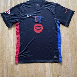 Svart FC Barcelona Nike fotbollströja - Snygg svart FC Barcelona fotbollströja från Nike med röda och blå detaljer längs sidorna. Tröjan har korta ärmar, klubbmärke på bröstet, Nike-logga och Spotify-tryck framtill. Tillverkad i lätt och ventilerande polyester, perfekt för match eller träning.