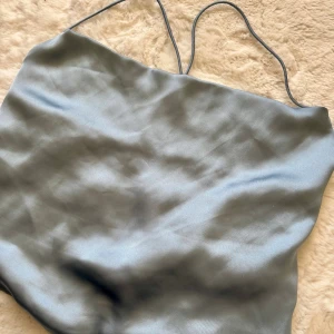 Ljusblå satinlinne från NA-KD - Supersnygg ljusblå linne-topp från NA-KD i satinliknande material. Tunt tyg med smala axelband och snygg knytning i ryggen. Perfekt för dig som gillar minimalistisk och stilren look. Passar till både byxor och kjol.