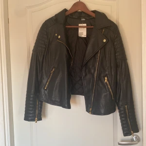 Svart skinnjacka från H&M, stl 40 - Säljer en svart skinnjacka från H&M i storlek 40. Jackan har coola quiltade detaljer på axlar och ärmar, guldiga dragkedjor och klassisk bikerkrage. Snygg croppad passform och markerade sömmar ger en edgy look. Perfekt för dig som vill sticka ut lite extra.
