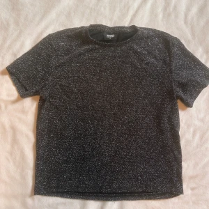 Svart glittrig t-shirt från DRDENIM - Svart t-shirt från DRDENIM med glittrigt mönster som ger en snygg shimmer-effekt. Modellen är klassisk med rund hals och korta ärmar. Perfekt för dig som vill sticka ut lite extra med en cool och trendig look.