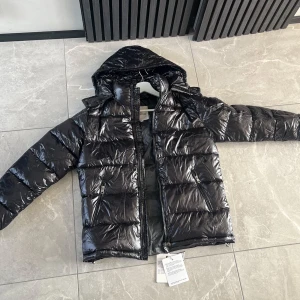 Svart Moncler pufferjacka med huva - Snygg svart pufferjacka från Moncler med glansig finish, huva och dragkedja framtill. Jackan har två dragkedjeförsedda fickor och ikonisk Moncler-logga på bröstet. Perfekt för kalla dagar när du vill hålla stilen.