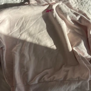 Ljusrosa hoodie från Reebok i M - Säljer en ljusrosa hoodie från Reebok i storlek M. Tröjan har en klassisk huva och är dekorerad med Reeboks logga broderad i rosa framtill. Mjuk och skön bomullsmix, perfekt för chill eller träning.