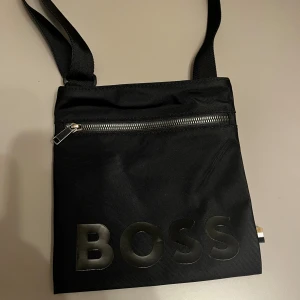 Svart axelväska från BOSS - Snygg svart axelväska från BOSS med justerbar rem och stort BOSS-tryck framtill. Väskan har en praktisk dragkedja upptill och är tillverkad i slitstark polyester. Perfekt storlek för mobil, plånbok och andra småsaker.