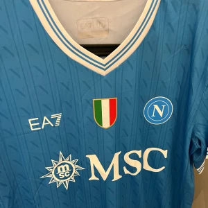 Napoli EA7 blå fotbollströja - Napoli fotbollströja från EA7 i blått med vita detaljer och klubbmärke på bröstet. Tröjan har V-ringning med vita och blå ränder, tryckta sponsorer och italienskt mästerskapsmärke. Tillverkad i lätt och ventilerande material, perfekt för match eller träning. 