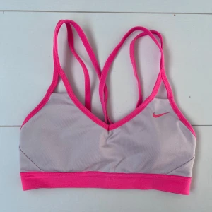 Nike Dri-Fit rosa sportlinnetopp XS - Nike Dri-Fit sportlinnetopp i ljusgrått med starkt rosa kant och axelband. Snygg v-ringad front och korsade smala band i ryggen. Perfekt för träning med fuktavvisande material och elastisk passform. Ikonisk Nike-logga framtill.