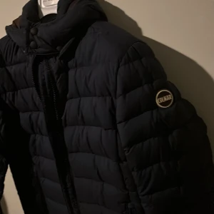 Mörkblå Colmar pufferjacka - Säljer en mörkblå pufferjacka från Colmar med klassisk logga på ärmen. Jackan har hög krage, dragkedja framtill och två sidofickor med dragkedja. Perfekt för kalla dagar och riktigt snygg till streetwear-stilen. Helt ny. 