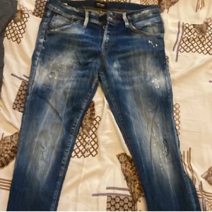 Blå jeans från Jack & Jones - Säljer ett par blå jeans från Jack & Jones med slitna detaljer och ljusa partier framtill och baktill. Jeansen har klassisk femficksdesign, normal passform och coola färgstänk för en trendig look. Perfekta för dig som gillar en avslappnad streetstil.