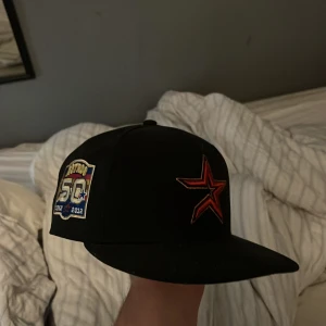 Svart New Era 59FIFTY Astros keps - Säljer en svart New Era 59FIFTY keps med Houston Astros röd stjärnlogga framtill och broderad 50th Anniversary-patch på sidan. Klassisk flat brim och Cooperstown Collection-märke. Tillverkad i 100% polyester.