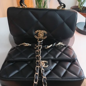 Svart quiltad handväska - Säljer en ikonisk svart handväska  med quiltad design och guldfärgade detaljer. Väskan har klassisk kedjerem med inslag av skinn och den välkända CC-loggan framtill. Perfekt för dig som vill ha en tidlös och lyxig accessoar.Båda för 270kr