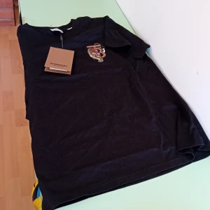 Svart Burberry t-shirt med tigerbrodyr - Svart t-shirt från Burberry i storlek XS med snygg broderad tiger och logga på bröstet. Klassisk passform och rund halsringning. Tillverkad i mjuk bomull, perfekt för en stilren och trendig look.