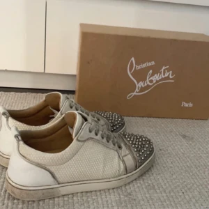 Christian Louboutin sneakers med nitar - Snygga vita sneakers från Christian Louboutin med ikoniska röda sulor och coola silverfärgade nitar på tån. Skorna har snörning och är tillverkade i skinn och textil med en unik struktur. Perfekta för dig som vill sticka ut med exklusiv streetstyle. Dessa är äkta & veriferade av Securo Fashion. Därav priset, Fått mycket komplimanger och sticker ut lite mer än de andra svarta ”fejk” loubsen kan gå ner i pris vid en snabb affär så kom med bud