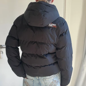 Svart pufferjacka från Superdry - Svart pufferjacka från Superdry med huva och dragkedja framtill. Jackan har broderad logga på bröstet och ärmen, samt två fickor med dragkedja. Perfekt för kalla dagar och har en sportig vibe.