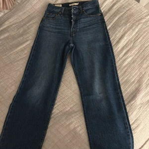 Levi's Ribcage Straight jeans blå W25 - Säljer ett par Levi's Premium Ribcage Straight jeans i klassisk blå tvätt. Hög midja, raka ben och snygga kontrastsömmar. Jeansen är tillverkade i bomull och har en tidlös look med fem fickor och knappgylf. Perfekta för dig som gillar en clean och stilren vibe.