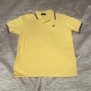 Gul pikétröja från Fred Perry - Snygg gul pikétröja från Fred Perry med klassisk krage och knäppning framtill. Tröjan har svarta och röda ränder på krage och ärmslut samt den ikoniska Fred Perry-loggan broderad på bröstet. Perfekt för en fräsch och sportig look.