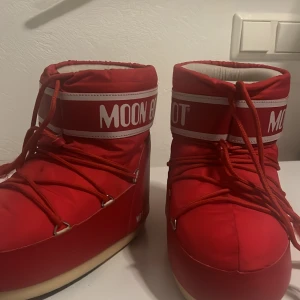 Röda Moon Boots med snörning - Säljer ett par röda Moon Boots med vit logga och snörning framtill. Skorna har en rund form, platt sula och är fodrade med mjukt syntetmaterial på insidan. Perfekta för vintern och tål kyla superbra! Snörena har slitits en del som man ser op bilderna, bara att skicka om man vill ha mer bilder eller har frågor mer funderingar!