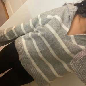 Gråvit randig stickad tröja V-ringad - Supermysig stickad tröja från minimarket i grått och vitt med breda ränder. V-ringad med krage och långa ärmar. Perfekt oversized look och ribbade muddar. Passar dig som gillar chill och stilren vibe.