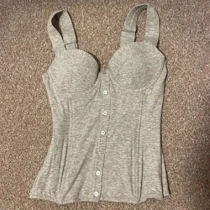 Grå korsettopp från Lindex - Snygg grå korsettopp från Lindex i nyskick. Den har knappar framtill och breda axelband för extra stöd. Baksidan har en smockdetalj som ger en bekväm passform. Perfekt för både vardag och fest! Storlek 75A