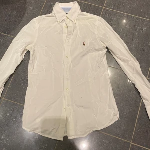 Vit skjorta från Ralph Lauren S - Vit långärmad skjorta från Ralph Lauren med klassisk button down-krage och broderad logga på bröstet. Skjortan är i bomull och har knappar framtill. Perfekt för en clean och stilren look. Obs, notera de två små fläckarna på sista bilden! 