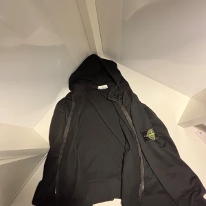 Svart hoodie från Stone Island - Svart hoodie från Stone Island med klassisk patch på vänster ärm. Tröjan har huva, dragkedja framtill och två fickor. Perfekt för dig som gillar streetwear och vill ha en clean look med snygga detaljer.