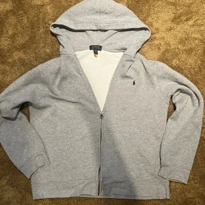 Grå hoodie från Polo Ralph Lauren - Säljer en grå hoodie från Polo Ralph Lauren med dragkedja och huva. Tröjan har långa ärmar, två fickor fram och den klassiska lilla Polo-loggan broderad på bröstet. Perfekt för chill dagar och enkel att matcha med jeans eller joggers.