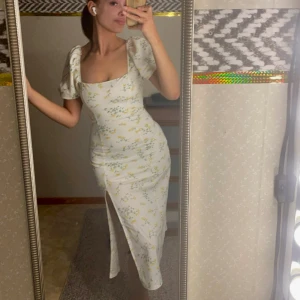 Blommig långklänning från Gina Tricot - Supersöt långklänning från Gina Tricot i vitt med gult och grönt blommönster. Klänningen har puffiga kortärmar, fyrkantig ringning och en snygg slits framtill. Perfekt för sommarens alla tillfällen! Endast använd två gånger 🌼