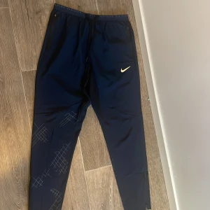 Nike Running Division träningsbyxor - Mörkblå träningsbyxor från Nike Running Division med smal passform, diskreta grafiska mönster på benen och dragkedja vid benslut. Byxorna har elastisk midja med snörning och praktiska fickor. Perfekta för löpning eller gymmet.