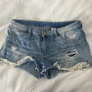 Ljusa jeansshorts med spets från &Denim - Snygga ljusblå jeansshorts från &Denim med slitna detaljer och hål framtill. Shortsen har råa kanter och vit spets längs bensluten för en extra cool touch. Klassisk femficksmodell med bälteshällor och knappstängning. Perfekta för sommaren!
