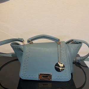 Blå axelväska med nitar från Alex Max - Snygg ljusblå axelväska från Alex Max med guldfärgade metalldetaljer. Väskan har både handtag och justerbar axelrem, samt praktiska fack inuti och dragkedja baktill. Perfekt för dig som vill ha en stilren men edgy look. Varan är i nyskick och aldrig använd💕Priset är diskuterbart!