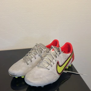 Nike Tiempo grå/röda fotbollsskor - Nike Tiempo fotbollsskor legend 9 i vitt och rött med neongula detaljer och svart kontur på swoosh-loggan. Skorna har snörning, rund tå och platt sula med dobbar. Insidan är röd och materialet är syntet med textur för bättre bollkontroll. Perfekta för dig som vill sticka ut på planen. Priset är diskuterbart och skorna rengörs noga innan leverans💕