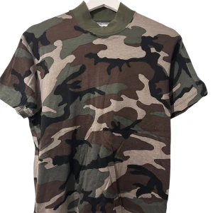 Camouflage t-shirt med grön krage - Säljer en kortärmad t-shirt från GENES med klassiskt camouflage-mönster i grönt, brunt, beige och svart. T-shirten har en bred, ribbad grön krage som ger en cool touch. Materialet är mjuk bomull.