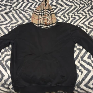 burberry hoodie - Äkta Svart hoodie från Burberry med klassisk beige, svart och röd rutig huva. Huva med snörning och stora fickor framtill. Perfekt för dig som vill ha en stilren men ändå lyxig streetwear-look.