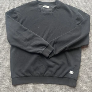 Svart sweatshirt från Jack & Jones - En stilren svart sweatshirt från Jack & Jones med rund halsringning och ribbade muddar. Tröjan är i mjuk bomullsmix och har en liten vit logolapp nertill. Perfekt för en chill och enkel look. Svarar ofta snabbt så hör av er vid frågor och funderingar🙌. kom gärna med prisförslag så kan vi säkert lösa något som funkar för båda😄