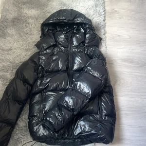Svart pufferjacka från Everest -  Snygg svart pufferjacka från Everest med glansig finish och huva. Jackan har dragkedja framtill, två fickor med dragkedja och en tydlig logga på ärmen. Perfekt för kalla dagar och riktigt trendig look.