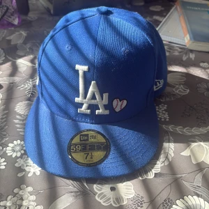 Blå LA Dodgers keps från New Era - Säljer en blå LA Dodgers 59FIFTY keps från New Era med broderade detaljer som LA-logga, ett hjärta med baseballsömmar, '1988 World Series'-patch och texten 'Love & Hustle = Game' på baksidan. Kepsen är i klassisk modell med böjd skärm och MLB-logga bak.