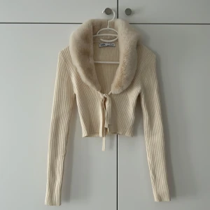 Beige ribbad tröja med fuskpäls från Zara - Superfin croppad tröja från Zara i beige ribbstickat material. Koftan har en bred, fluffig fuskpäls-krage och knytning framtill. Långärmad och perfekt för höst och vinter när du vill ha något extra mysigt och trendigt. Pris 350kr, ordinarie 599kr